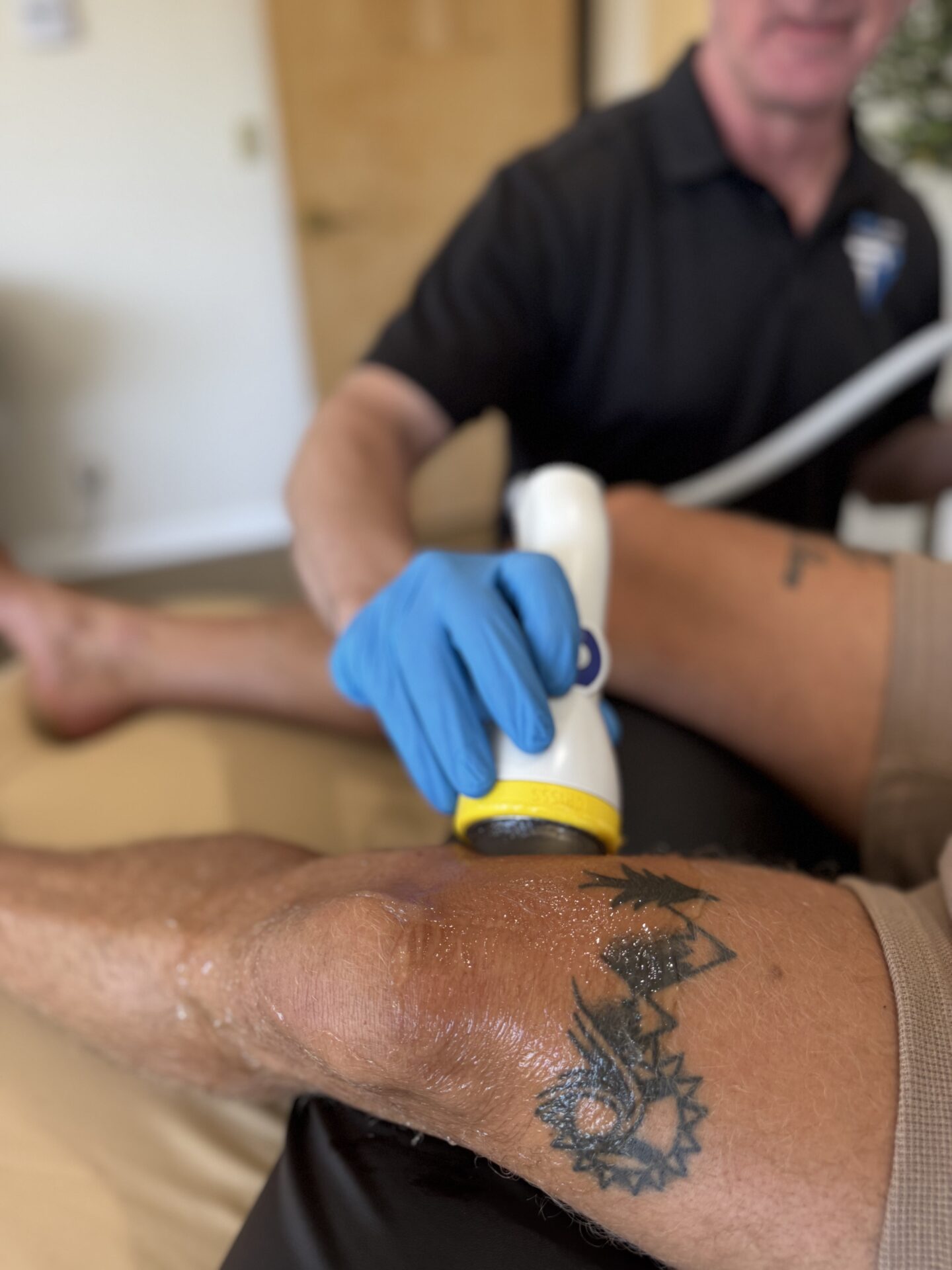 A cozy spa room with light blue brick walls. A massage table with white sheets stands near a wooden console table with decorative baskets and a clock. Two wooden shelves with various products and decor are on the wall. A beige chair with a blue cushion is beside the table.
